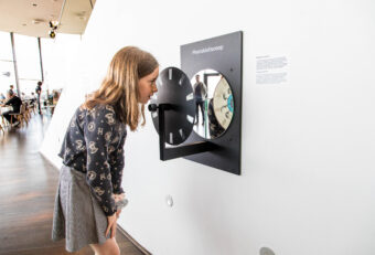 A woman in a floral top and gray skirt leans toward a wall-mounted interactive film reel exhibit in a bright museum gallery with white walls