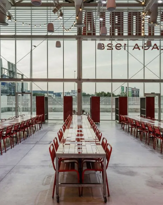 Largos pasillos de mesas blancas con sillas de metal rojas dentro de una luminosa nave industrial con paredes de cristal de suelo a techo que muestran el nombre Fábrica Cervezas Victoria en letras espejadas.