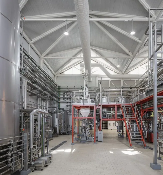 Interior de una cervecería con grandes tanques de fermentación de acero inoxidable, tuberías extensas, techo de cerchas blancas y plataformas de metal rojo con escaleras.