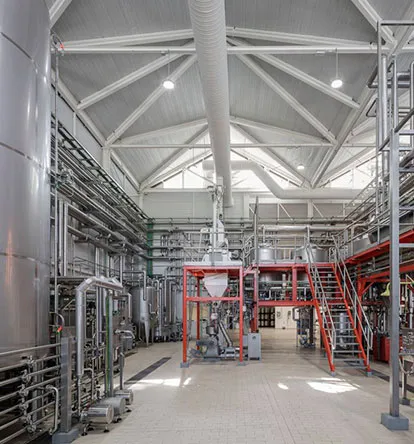 Interior de una planta de producción de cerveza con grandes tanques de acero inoxidable, tuberías extensas, escaleras y plataformas de metal rojo, y un techo alto con vigas blancas.