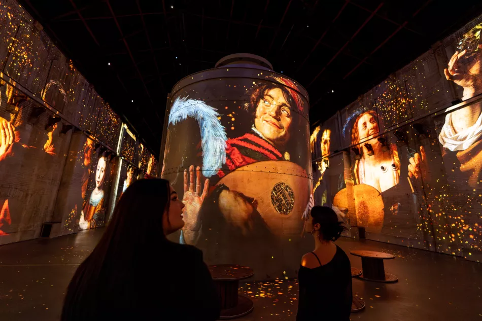 Immersive digital art exhibition with large projections of classical paintings on walls and cylindrical display, visitors observing in dark space
