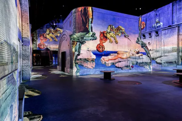 Proyecciones inmersivas de arte digital cubren las paredes de un espacio de exhibición tenuemente iluminado con arcos de piedra y texturas industriales.
