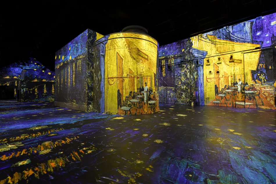 Inmersivas proyecciones de arte digital que cubren paredes y suelo en un espacio de exposición oscuro con imágenes coloridas en movimiento.