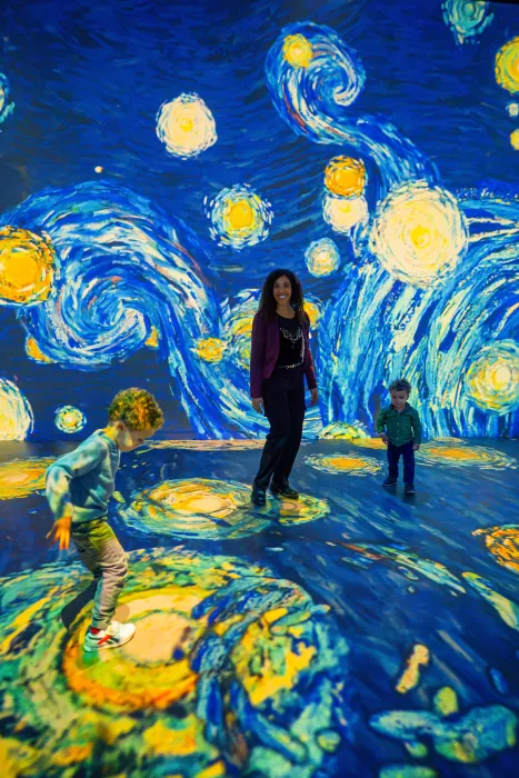 Los visitantes interactúan con proyecciones inmersivas de patrones arremolinados en azul y amarillo que se asemejan a La noche estrellada de Van Gogh en una sala oscura.