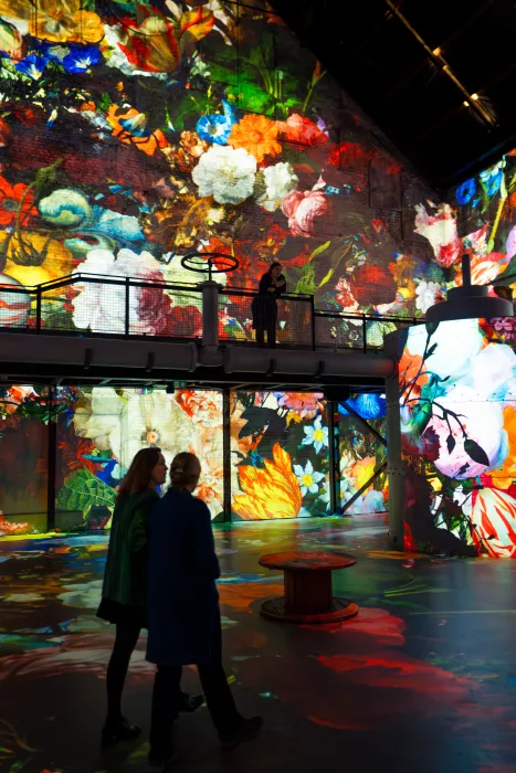 Immersive digital art installation with vibrant floral projections covering walls and floor; visitors walk through the space while one person stands on an elevated platform.