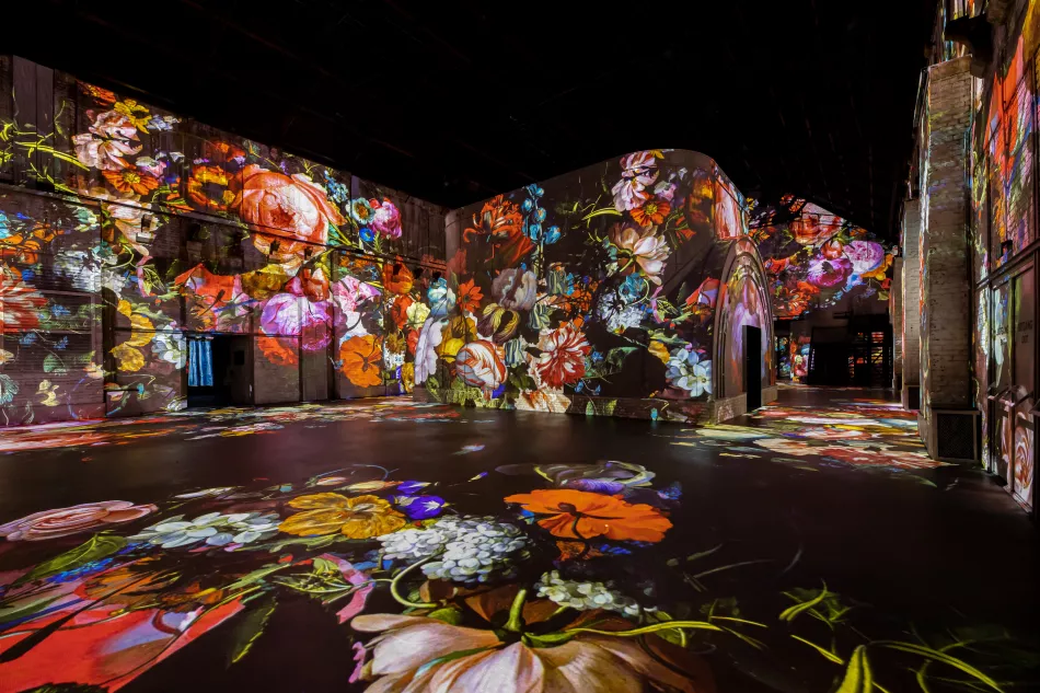 Proyecciones digitales inmersivas de vibrantes flores y arte clásico cubren las paredes y el suelo de una sala oscura en Fabrique des Lumières