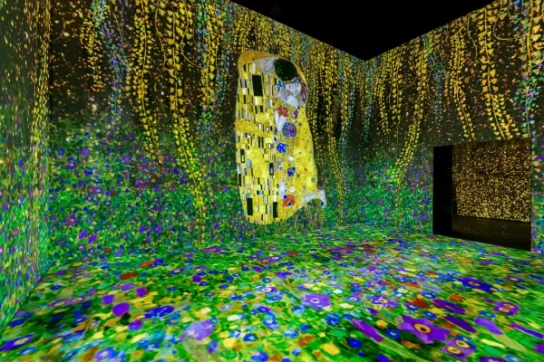 Proyección de arte digital inmersiva con vibrantes patrones florales que cubren paredes y suelo, con una obra de arte dorada central con figuras rodeadas de efectos de luz en cascada