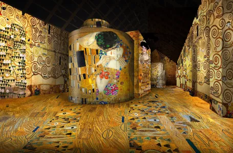 Proyecciones digitales inmersivas de patrones arremolinados dorados y figuras cubren paredes, suelo y un cilindro central en una sala de exposiciones oscura