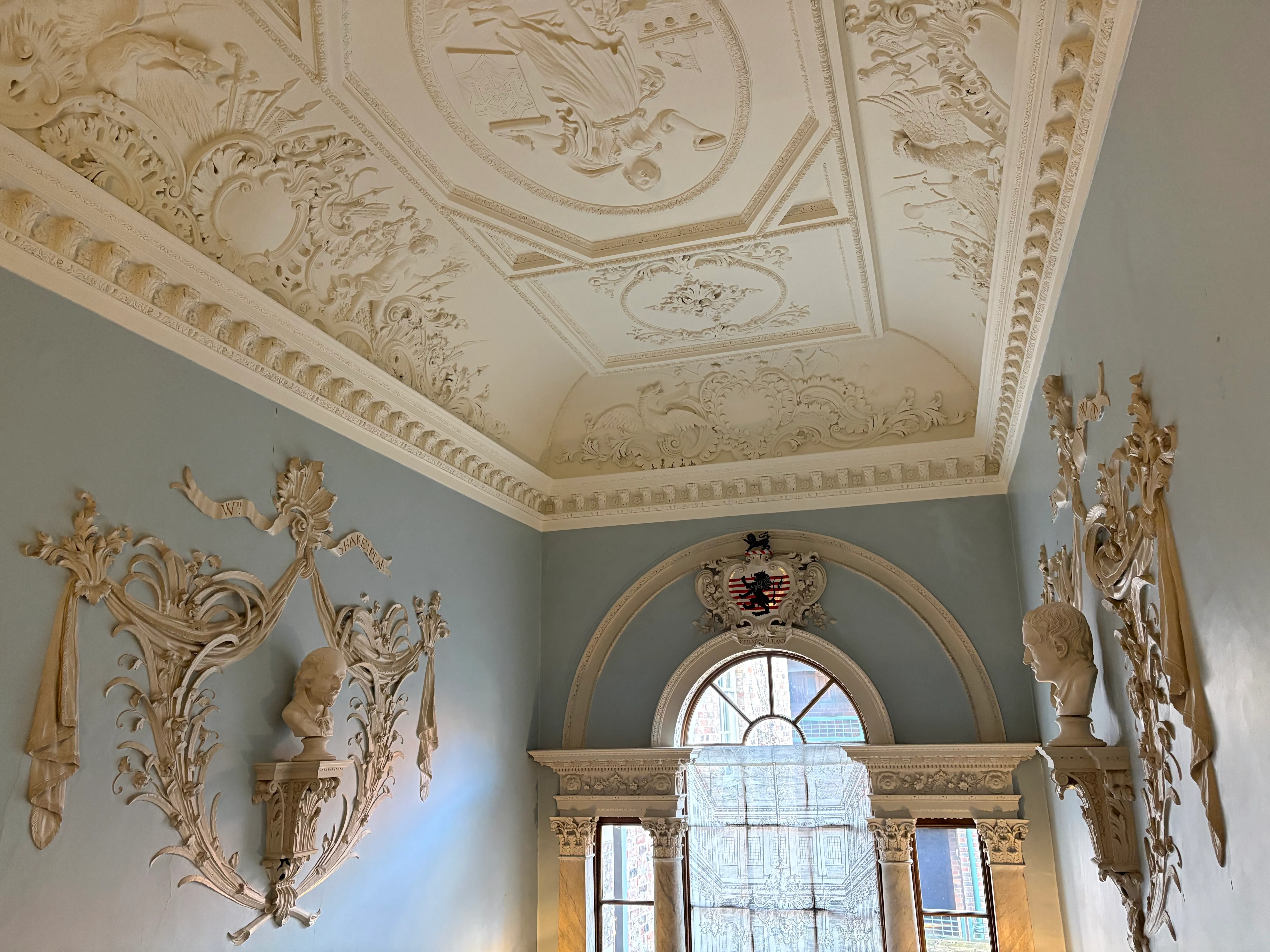 Ornado techo de yesería blanca con elaborados motivos decorativos sobre paredes de color gris azulado, adornadas con apliques dorados y una ventana arqueada al fondo de un gran salón