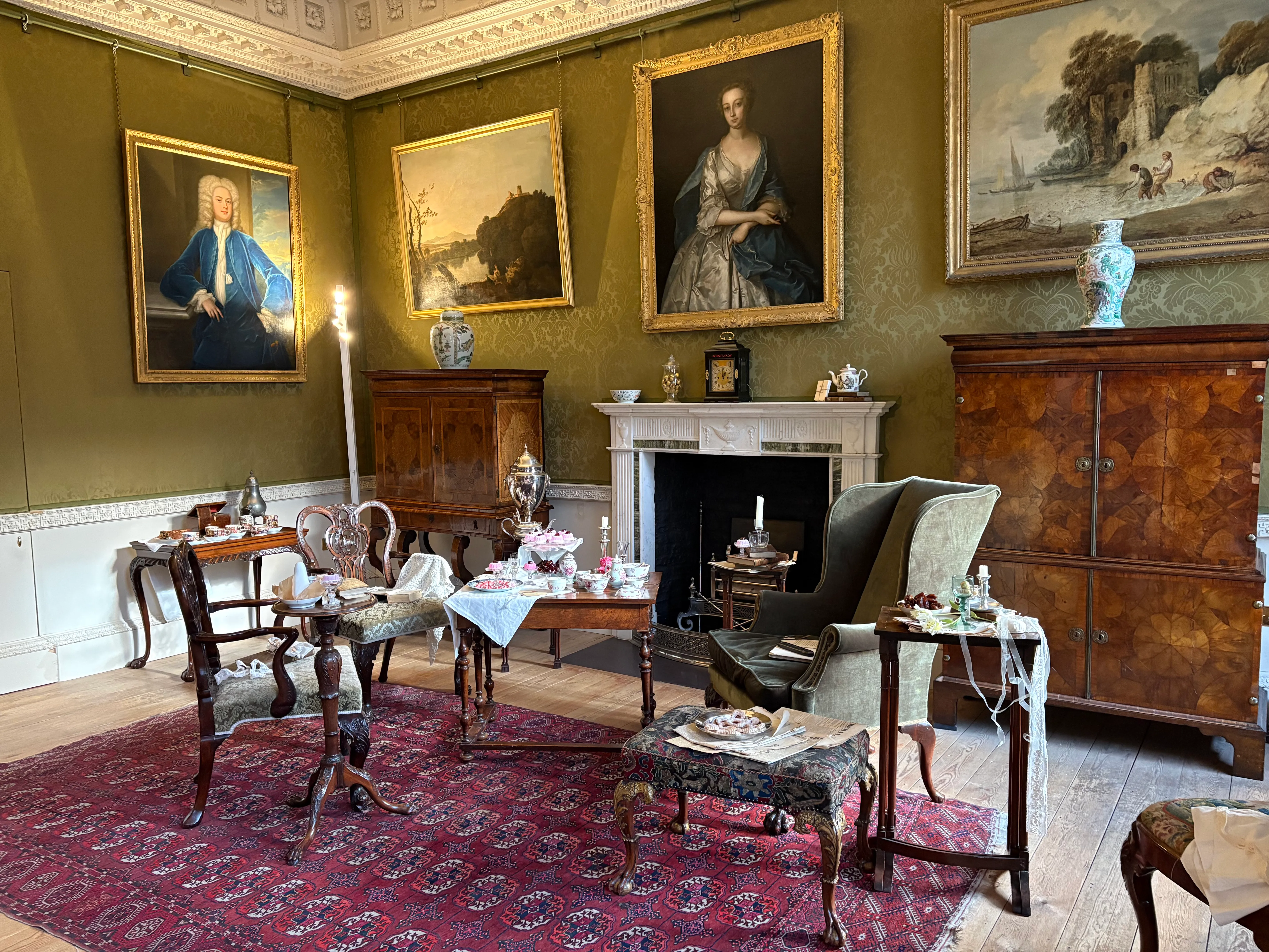 Un salón de estilo georgiano ricamente decorado con paredes de color verde oliva, grandes retratos en marcos dorados, una chimenea de mármol blanco y muebles de época dispuestos sobre una gran alfombra roja estampada.