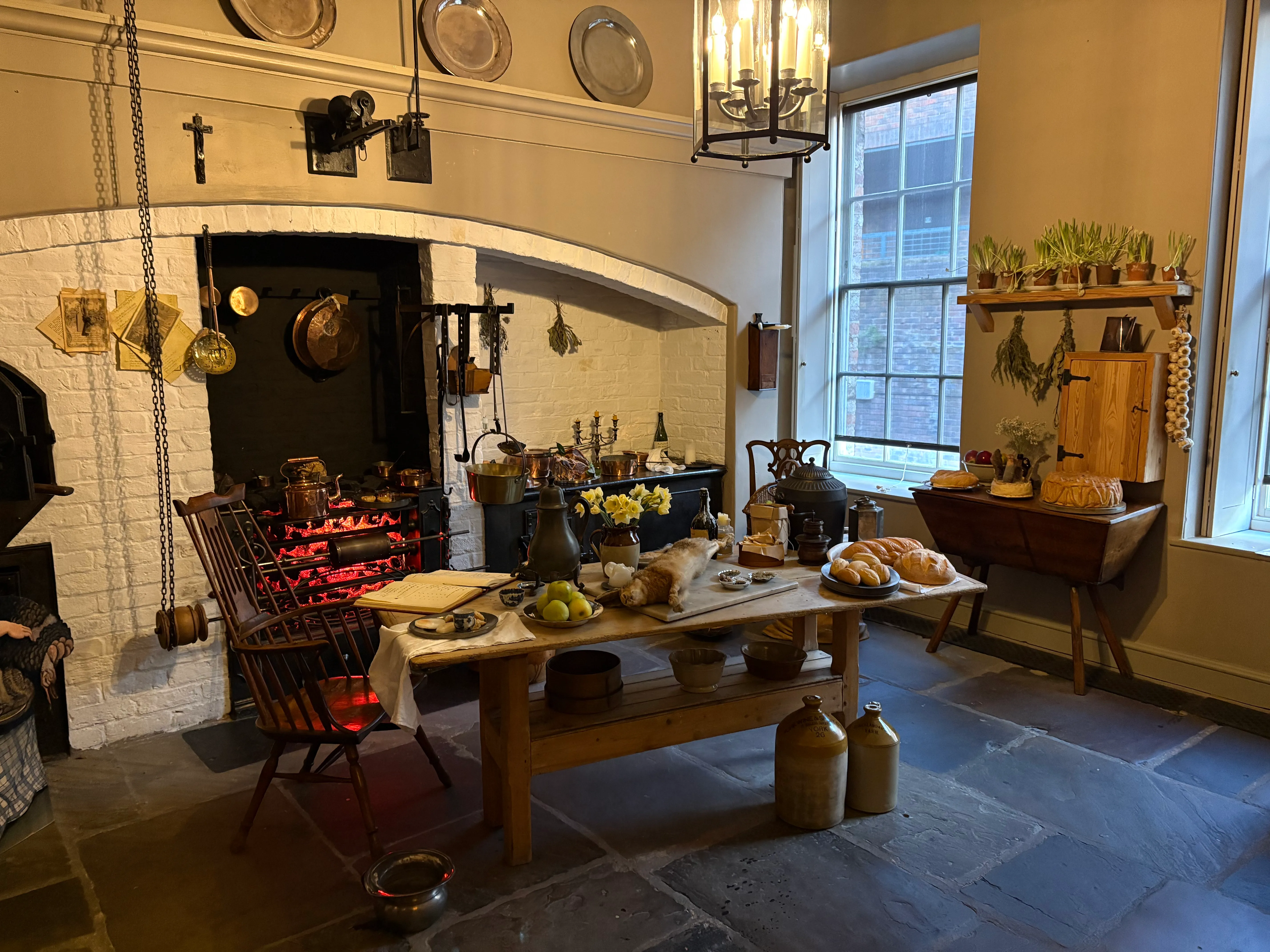 Interior de una cocina georgiana con una gran chimenea de ladrillo blanco, ollas y sartenes de cobre colgantes, una mesa de madera cargada de pan y fruta, y un suelo de losas de piedra.