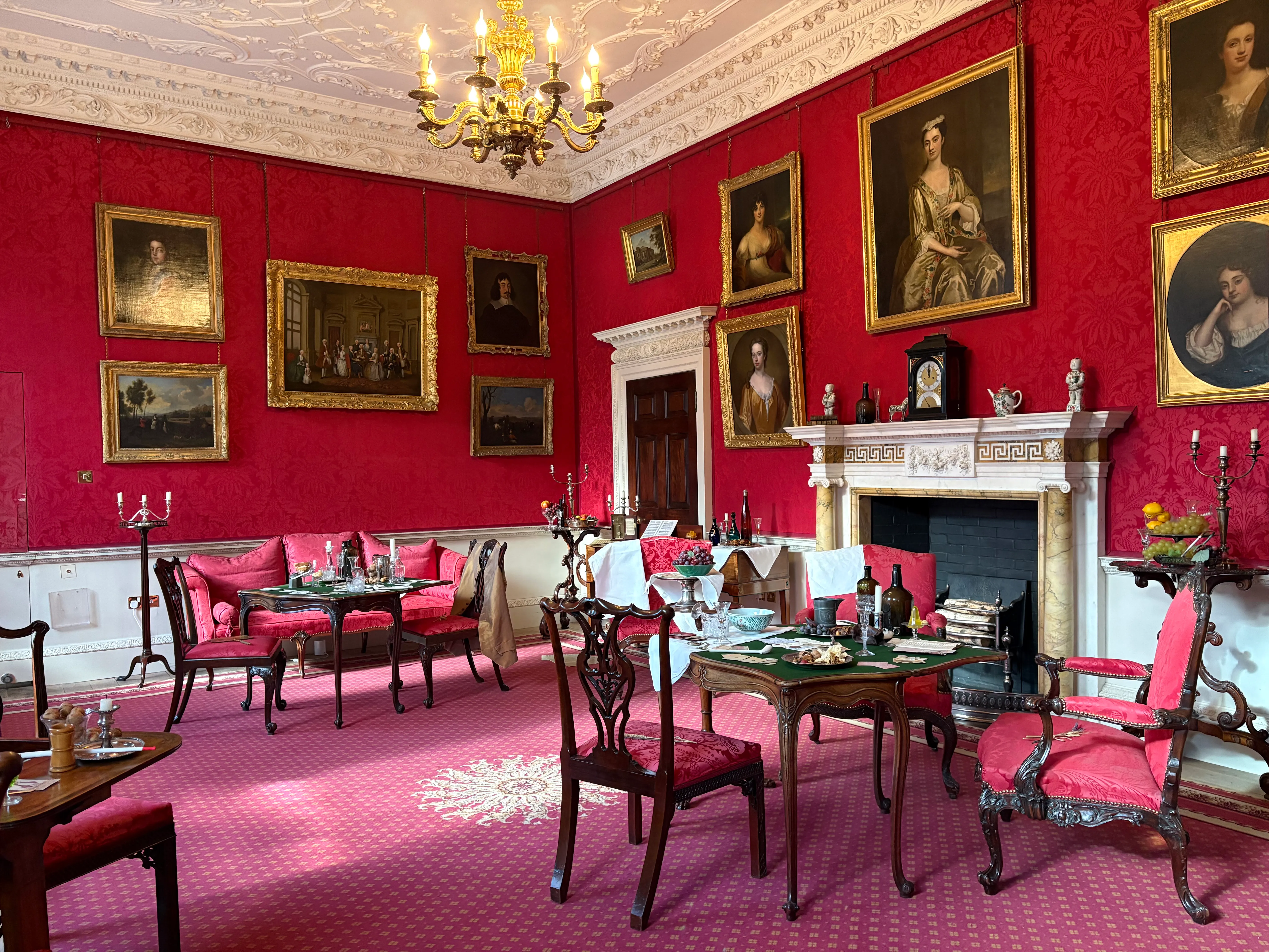 Salón georgiano ricamente decorado con paredes de color rojo intenso cubiertas de retratos enmarcados, chimenea de mármol blanco, lámpara de araña dorada y sillas y mesas de época dispuestas sobre una alfombra roja.