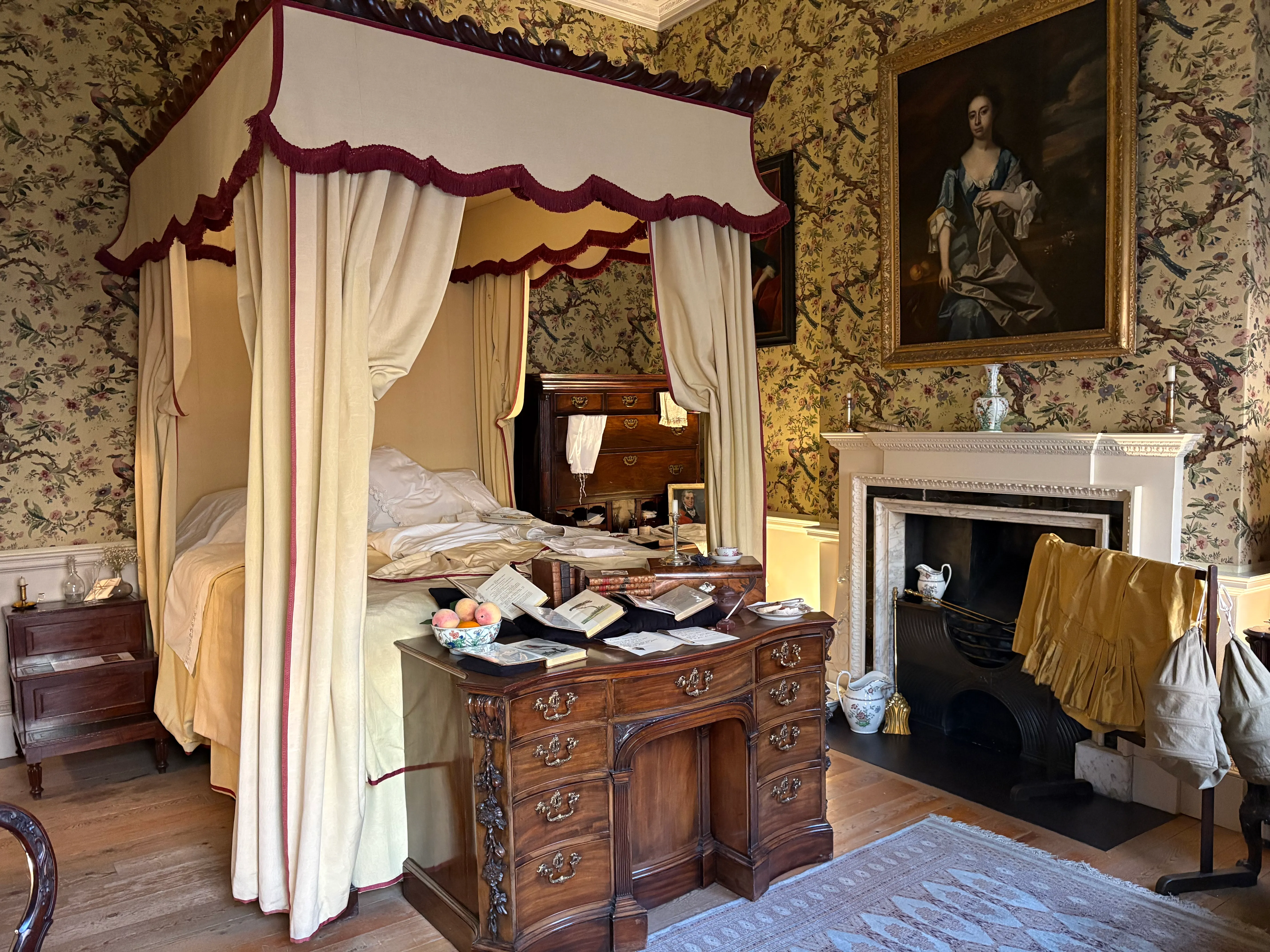 Interior de un dormitorio georgiano con una cama con dosel cubierta por telas de color crema y burdeos, un escritorio de madera y una chimenea con un retrato encima.