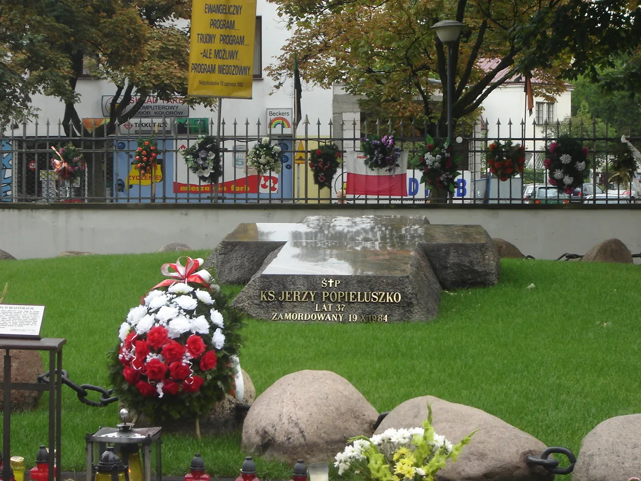 Een herinneringsgraf met een stenen markering met de inscriptie 'KS JERZY POPIEŁUSZKO', omringd door rode en witte bloemen, groen gras en kransen achter een hek.