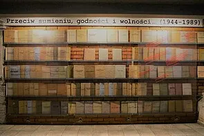 Una pared de estanterías llena de libros y documentos, con un letrero encima en polaco que dice 'Przeciw sumieniu, godności i wolności [1944-1989]'
