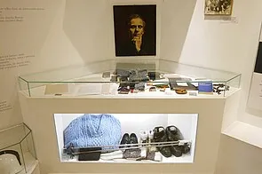 Glazen vitrine met persoonlijke spullen, waaronder een blauw kledingstuk, schoenen en een portret van een man aan de muur erboven