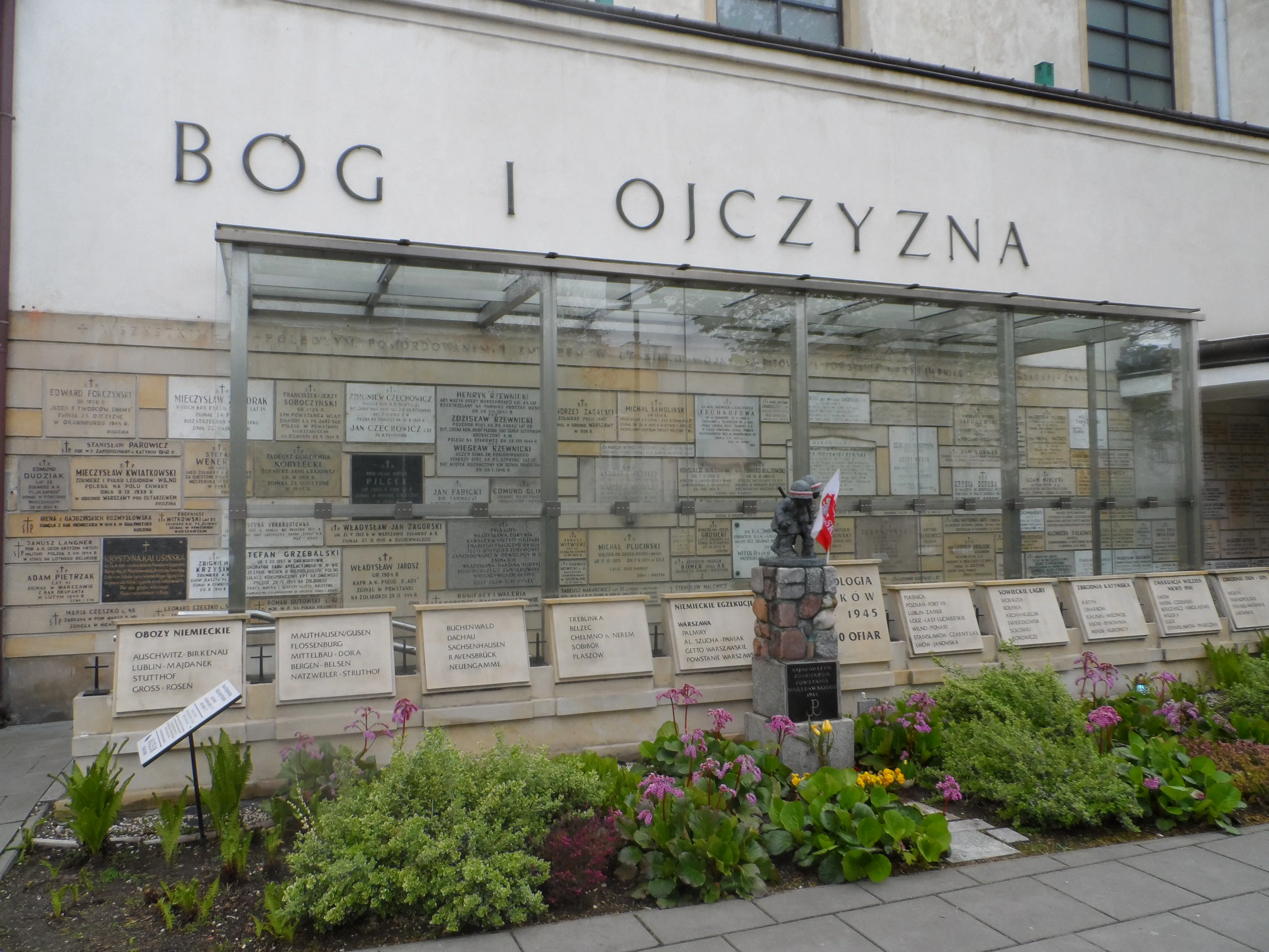 Herdenkingsmuur met plaquettes en de inscriptie 'BOG I OJCZYZNA' boven een glazen omheining, omringd door planten en bloemen