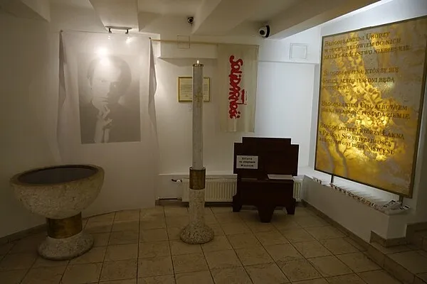 Interieur van een museum met een doopvont, een lange staf, een lessenaar en een groot verlicht display met tekst.