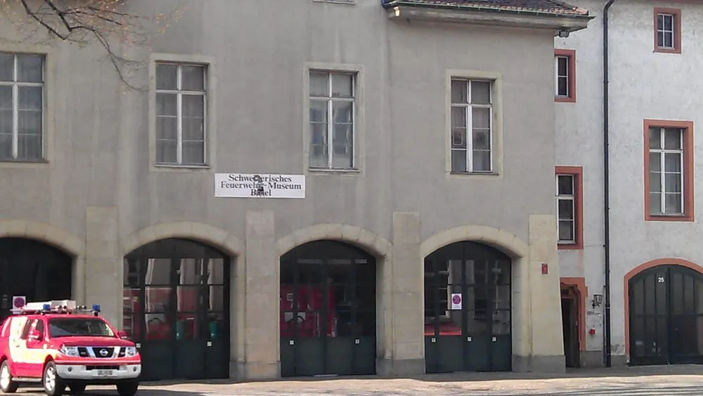 Fachada gris del Feuerwehrmuseum Basel con puertas de garaje arqueadas, un letrero que dice 'Feuerwehrmuseum' y un vehículo de bomberos rojo y blanco estacionado en frente.