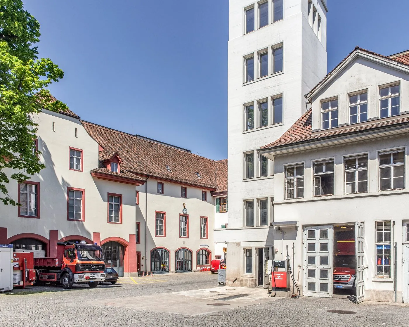 Patio del Feuerwehrmuseum Basel con edificios históricos blancos, bahías de garaje arqueadas con bordes rojos que albergan camiones de bomberos y una torre blanca alta bajo un cielo azul