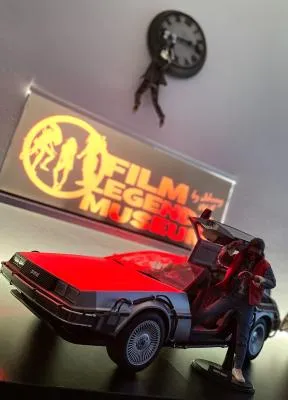 Una maqueta roja y negra del coche del tiempo DeLorean con una figura a su lado, delante de un letrero brillante del 'Film Legends Museum'.