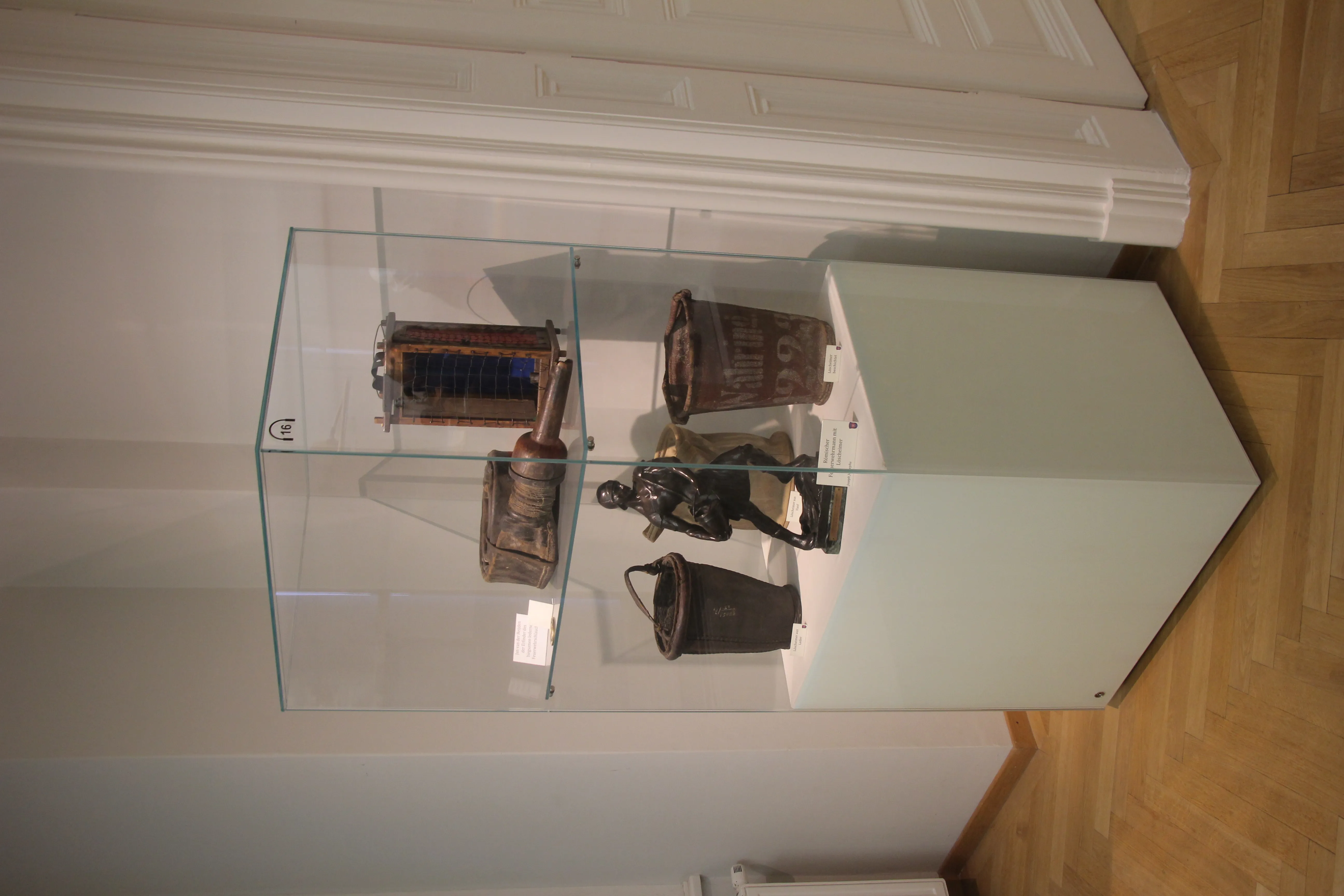 Glass display case containing historical firefighting equipment on a white pedestal in a museum gallery