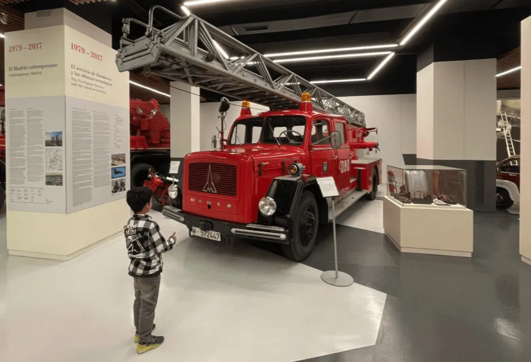 Un niño con camisa a cuadros está parado frente a un camión de bomberos vintage rojo dentro de una exhibición de museo.