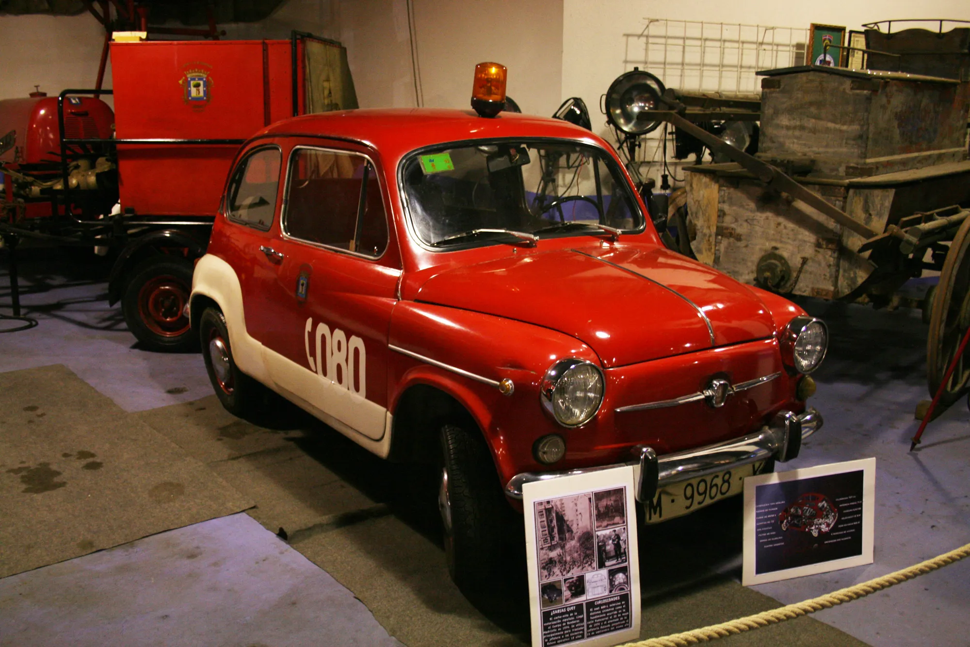 Un camión de bomberos vintage rojo y blanco con el texto 'BOM' en el lateral, exhibido en interiores con placas informativas y otro equipo al fondo.