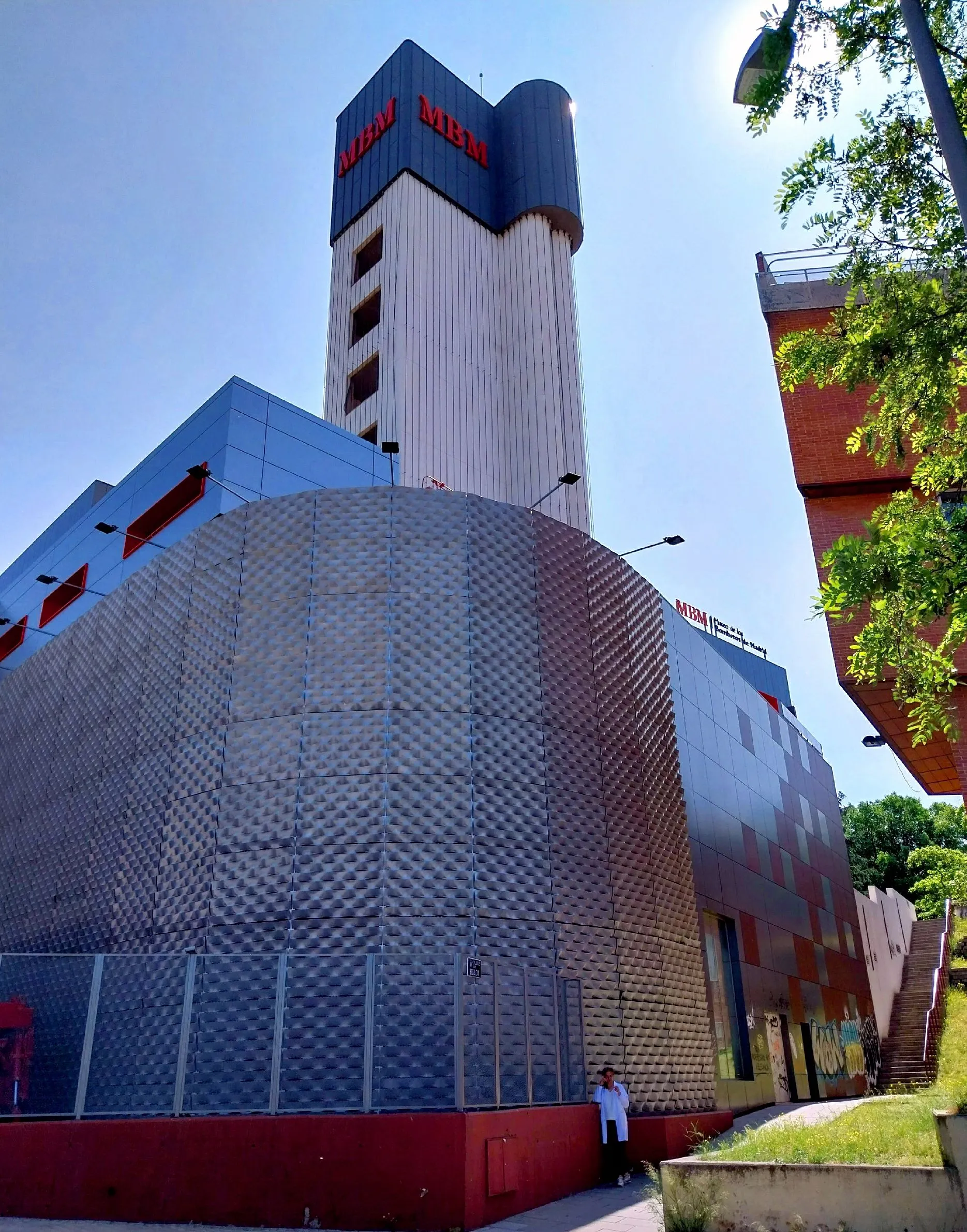 edificio moderno con una fachada metálica texturizada y una torre alta con letras rojas bajo un cielo azul claro