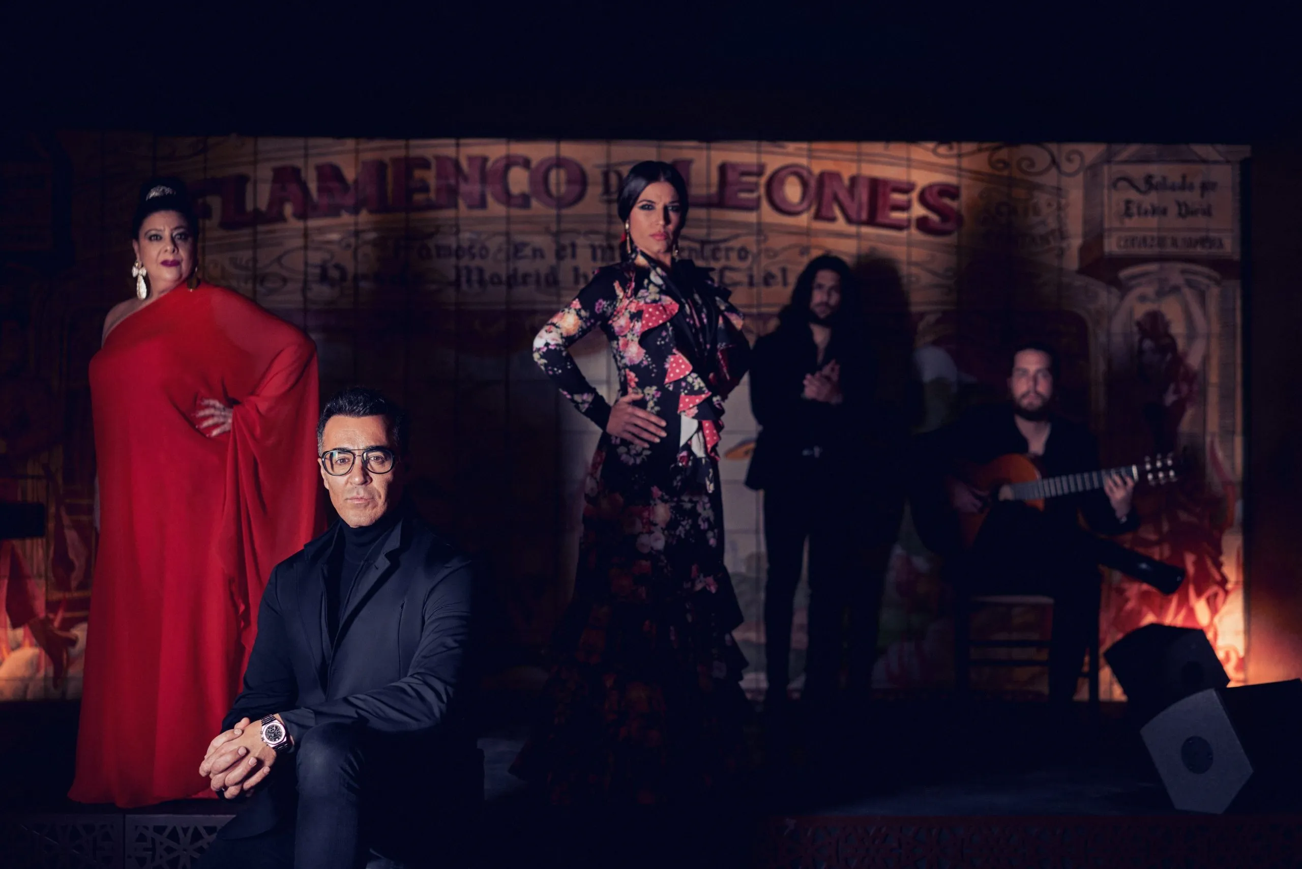 Cinco artistas de flamenco, incluyendo una mujer con vestido rojo, una mujer con vestido floreado y un hombre tocando la guitarra, de pie frente a un letrero de 'Flamenco de Leones'
