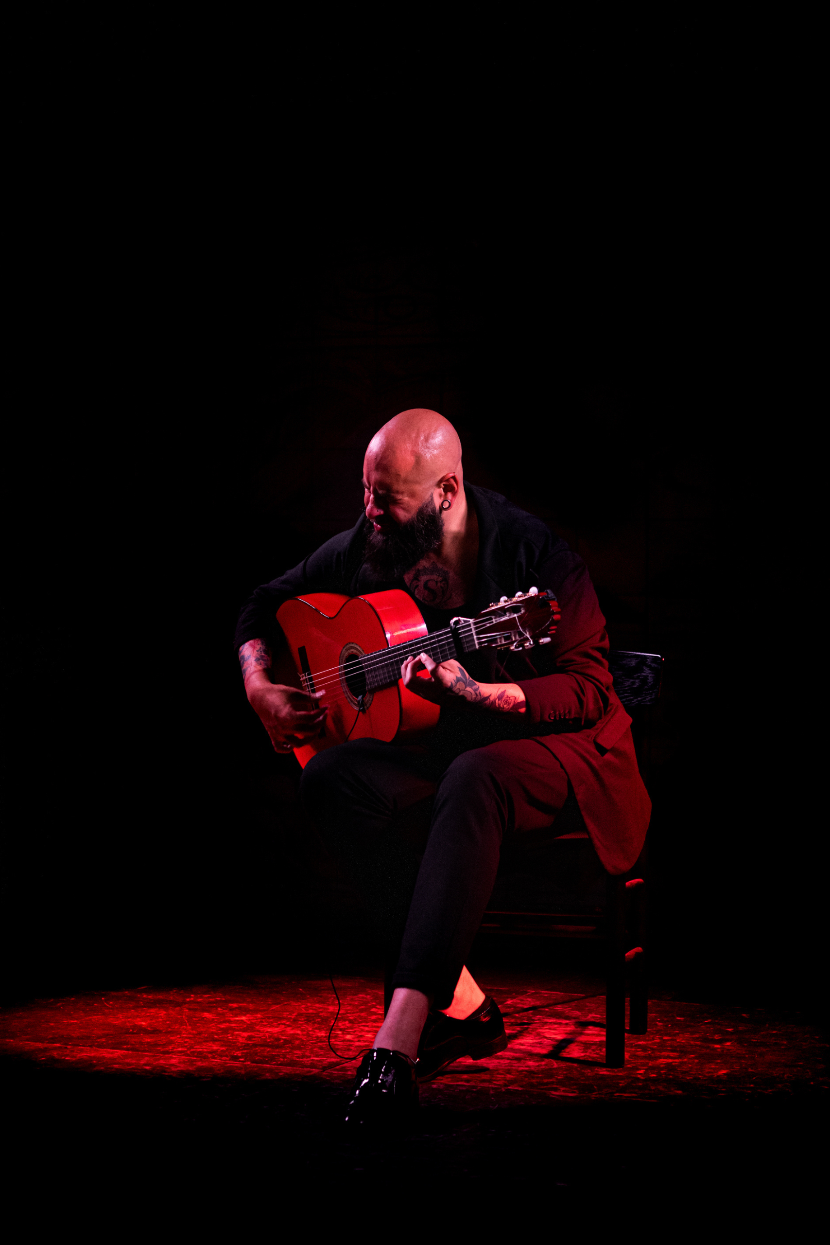 Un hombre calvo con barba tocando una guitarra clásica roja en un escenario oscuro iluminado por luz roja