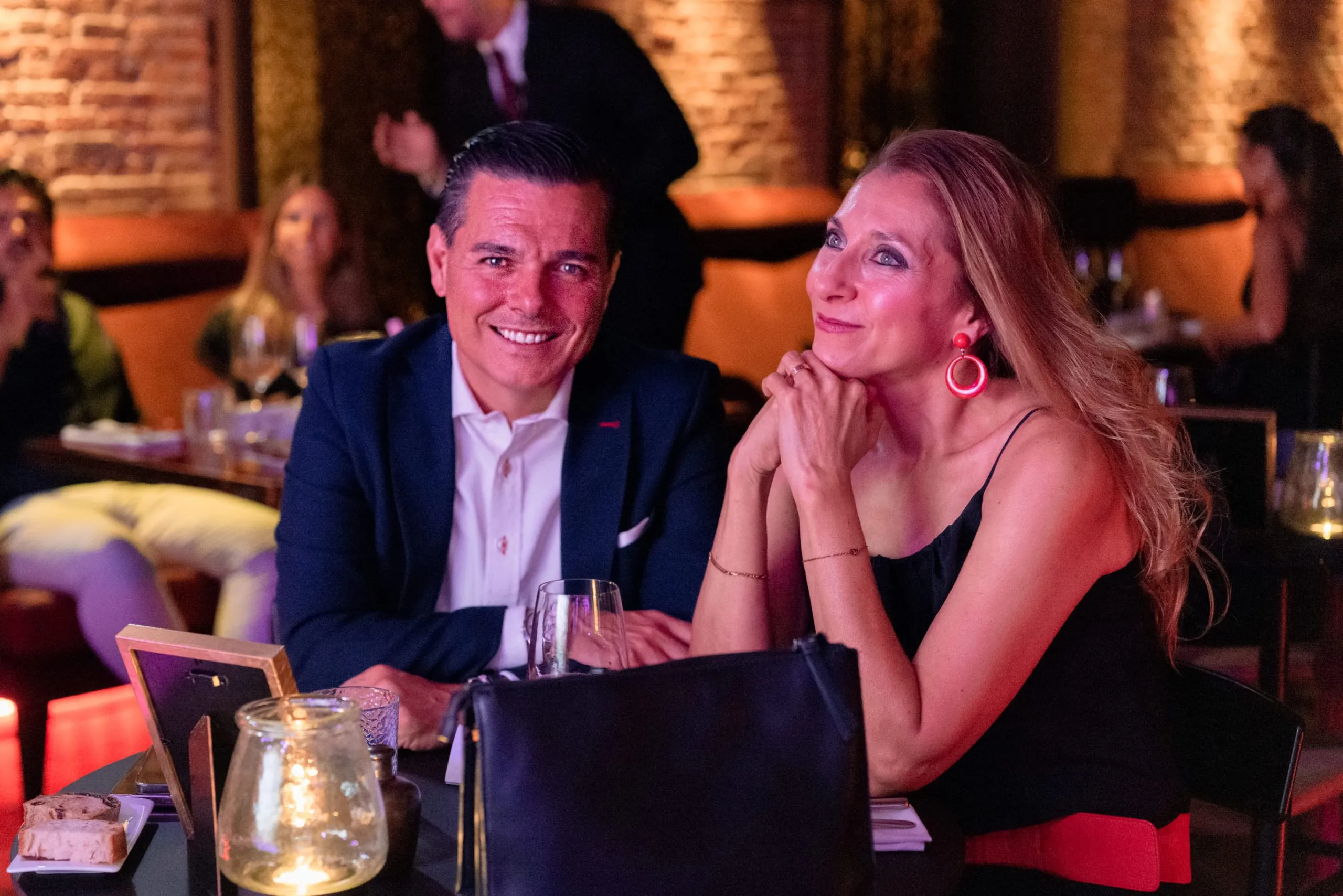 Mężczyzna i kobieta siedzą przy stoliku w słabo oświetlonej restauracji, uśmiechają się i patrzą w stronę sceny.