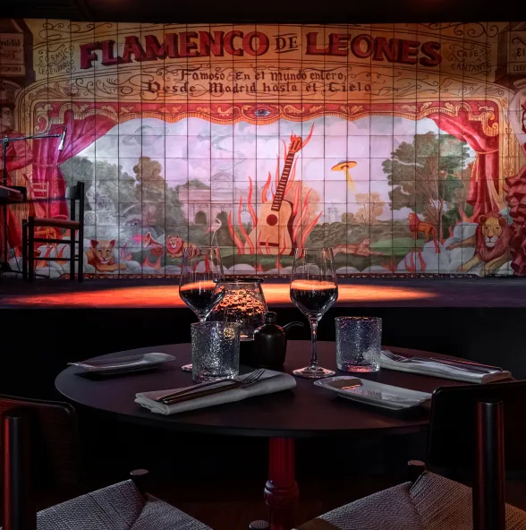 Una mesa redonda puesta para cenar con copas de vino y platos, frente a un escenario con un gran mural que dice 'Flamenco de Leones' y una guitarra pintada.