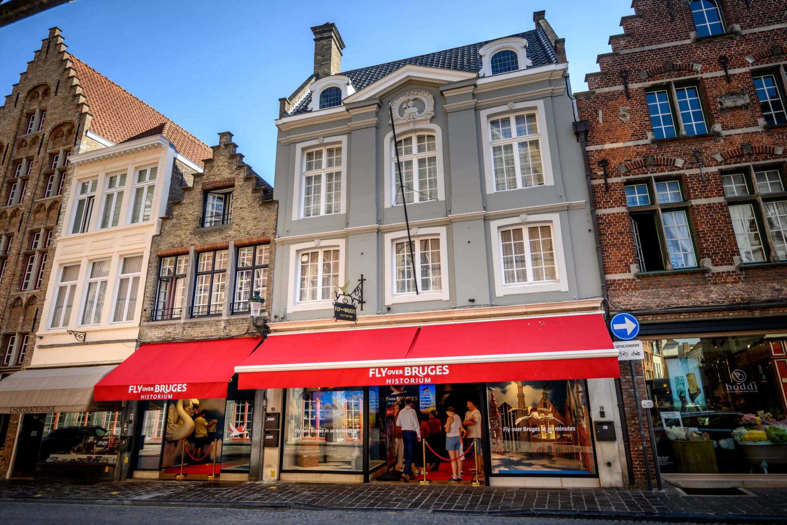 Vista de la calle de Fly Over Bruges con un toldo rojo brillante que cubre la fachada, flanqueado por edificios tradicionales de ladrillo con fachadas escalonadas bajo un cielo azul.