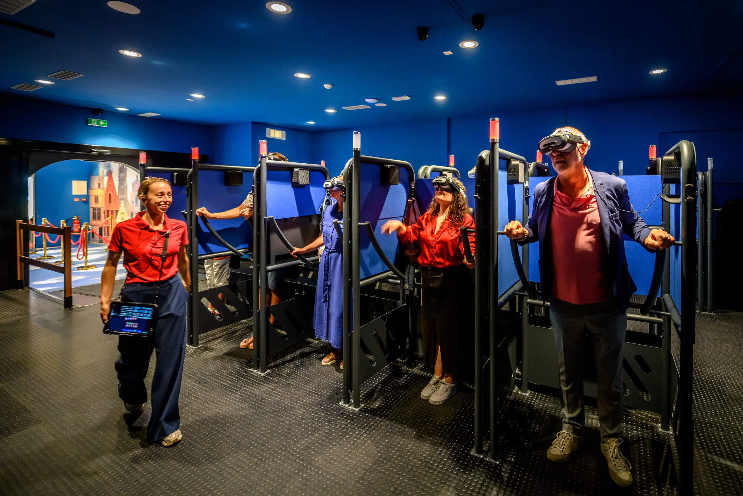 Personas con gafas de realidad virtual están de pie en cabinas individuales de simuladores de vuelo dentro de una sala de paredes azules, con un miembro del personal vestido con una camisa roja caminando cerca.
