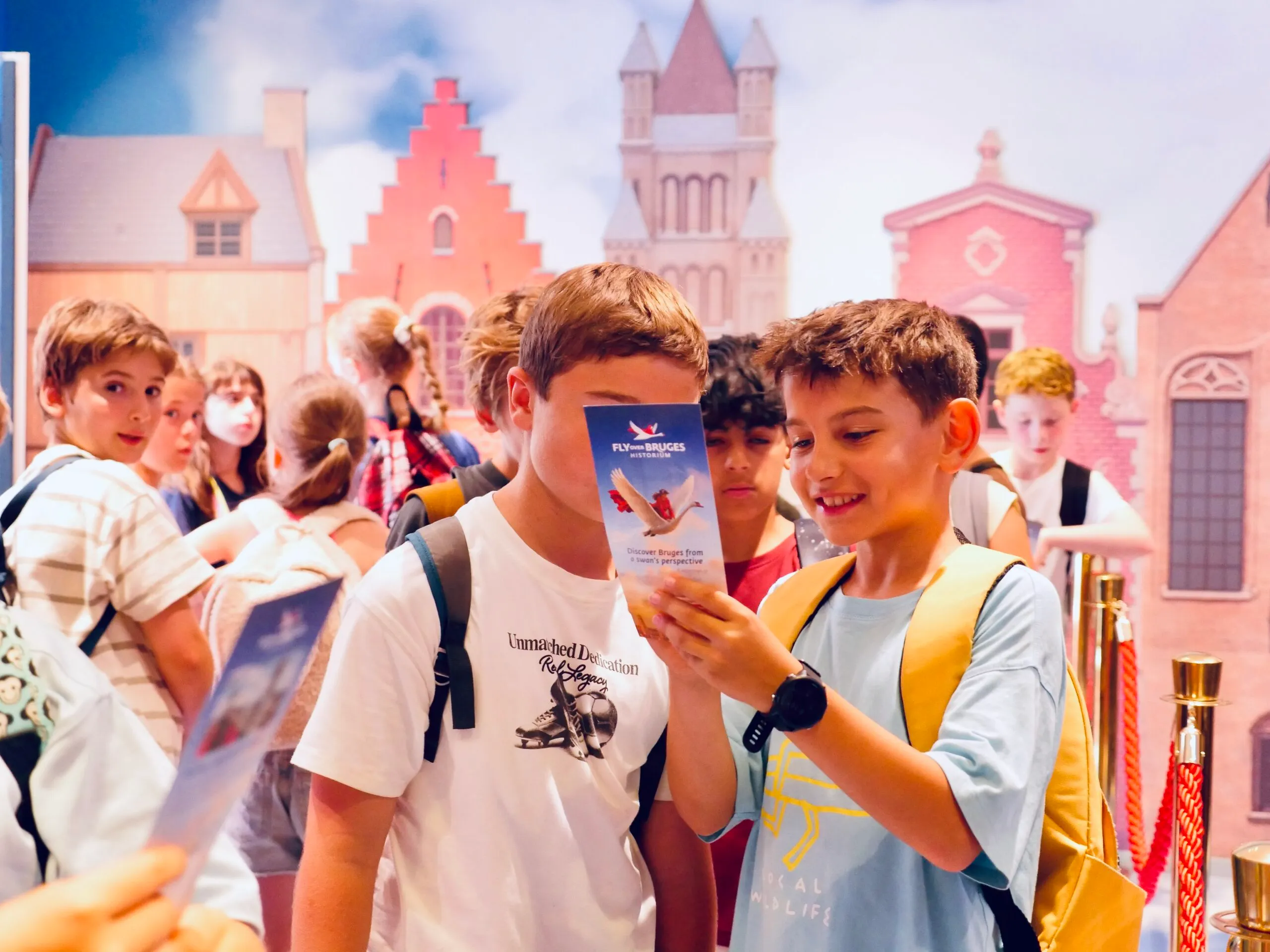 Dos chicos leyendo un folleto frente a un colorido telón de fondo de edificios de Brujas, con otros visitantes esperando en una zona de cola.