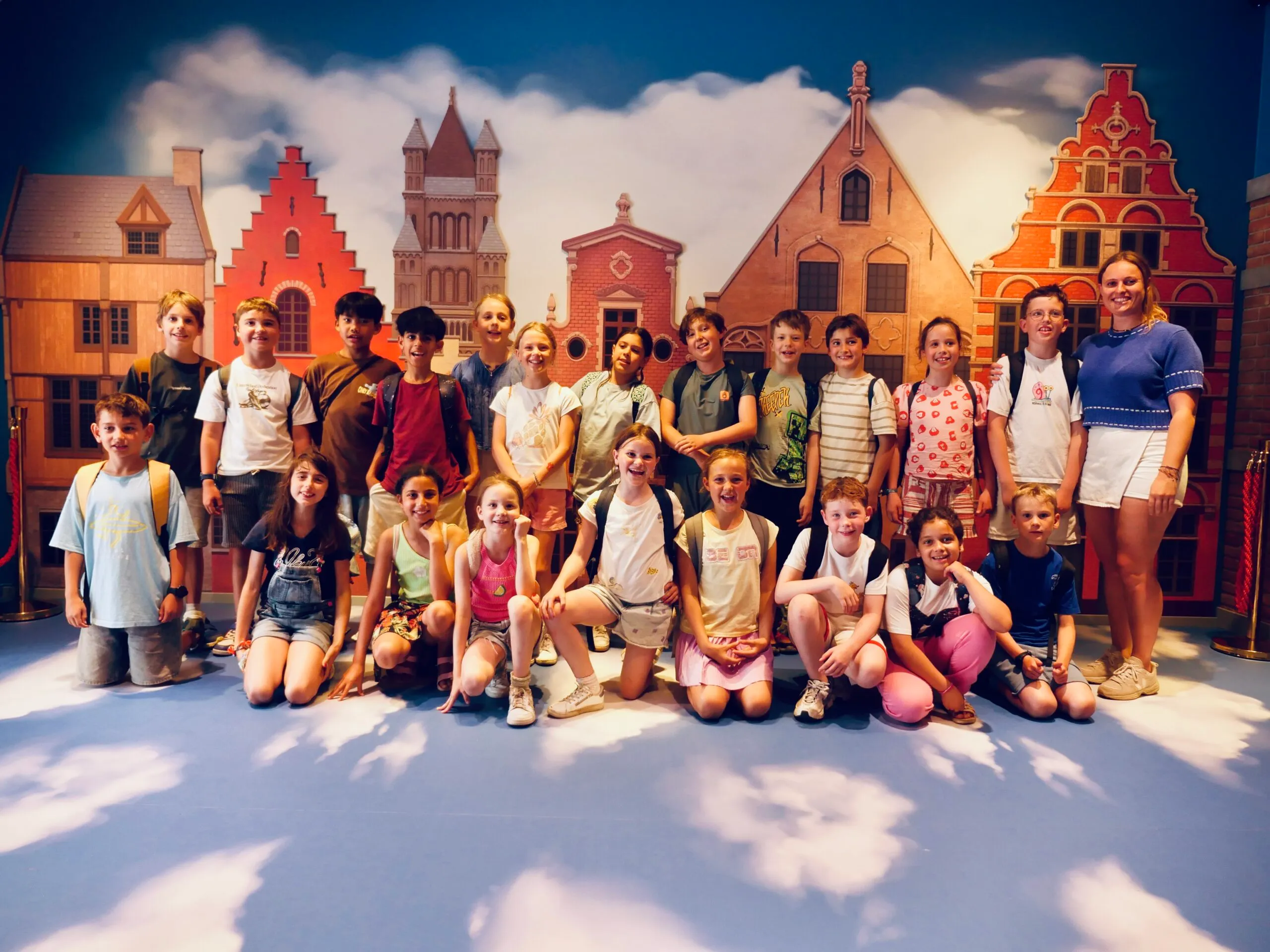 Grupo de niños y adultos posando frente a un mural colorido que representa edificios tradicionales de estilo flamenco con un cielo azul de fondo.