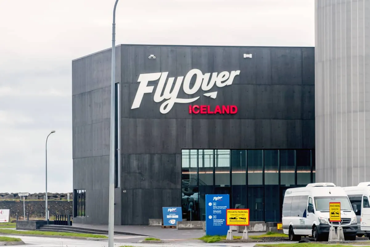 Vista exterior de un edificio gris oscuro con un gran cartel blanco y rojo de 'FlyOver Iceland' sobre la entrada de cristal, con furgonetas blancas aparcadas a la derecha.
