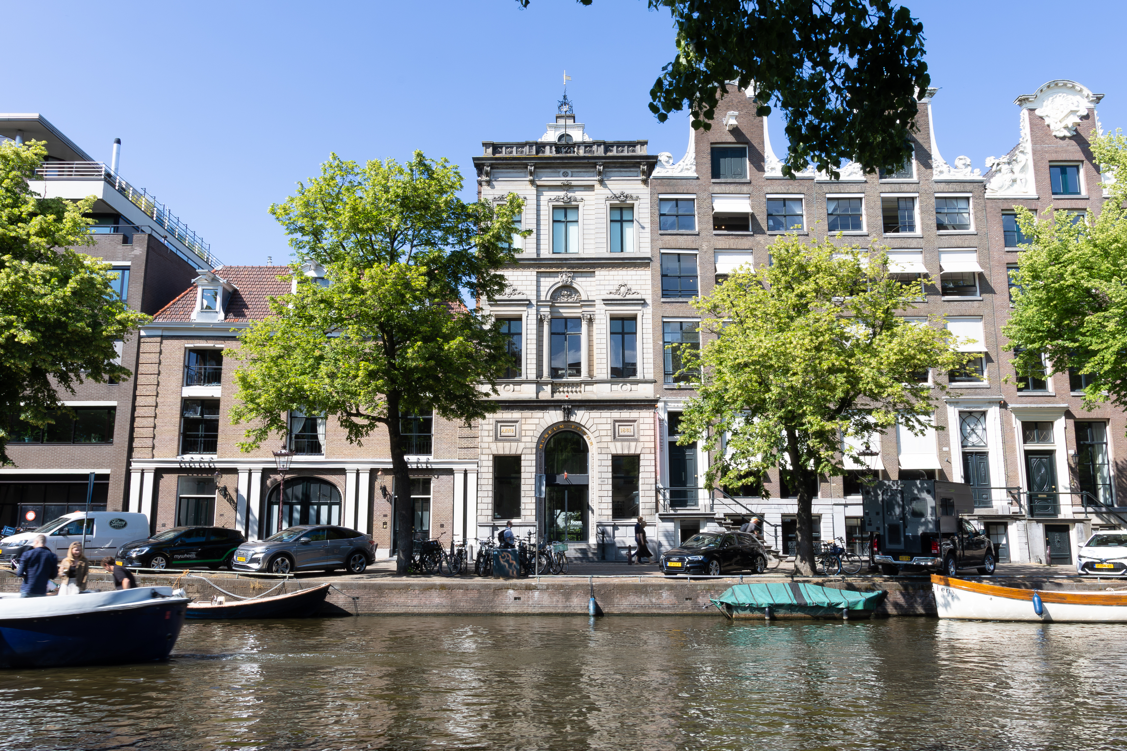 Rij van historische Nederlandse grachtenpanden met trapgevels langs een waterweg, aangemeerde boten, bomen die schaduw bieden op een zonnige dag