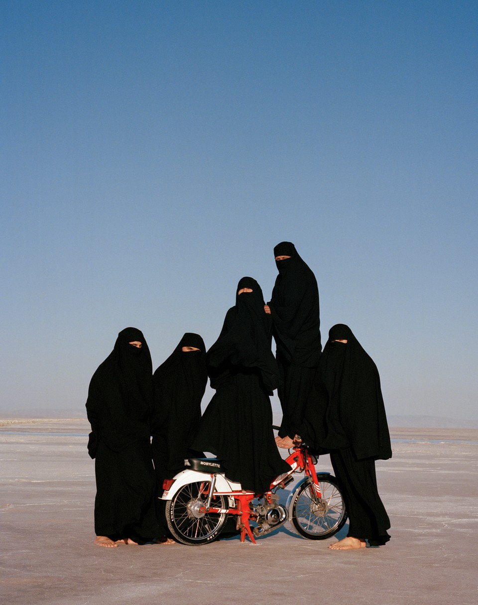 Zes mensen in zwarte abaya's verzamelden zich rond een rode en witte fiets in een uitgestrekt, vlak woestijnlandschap onder een helderblauwe hemel.