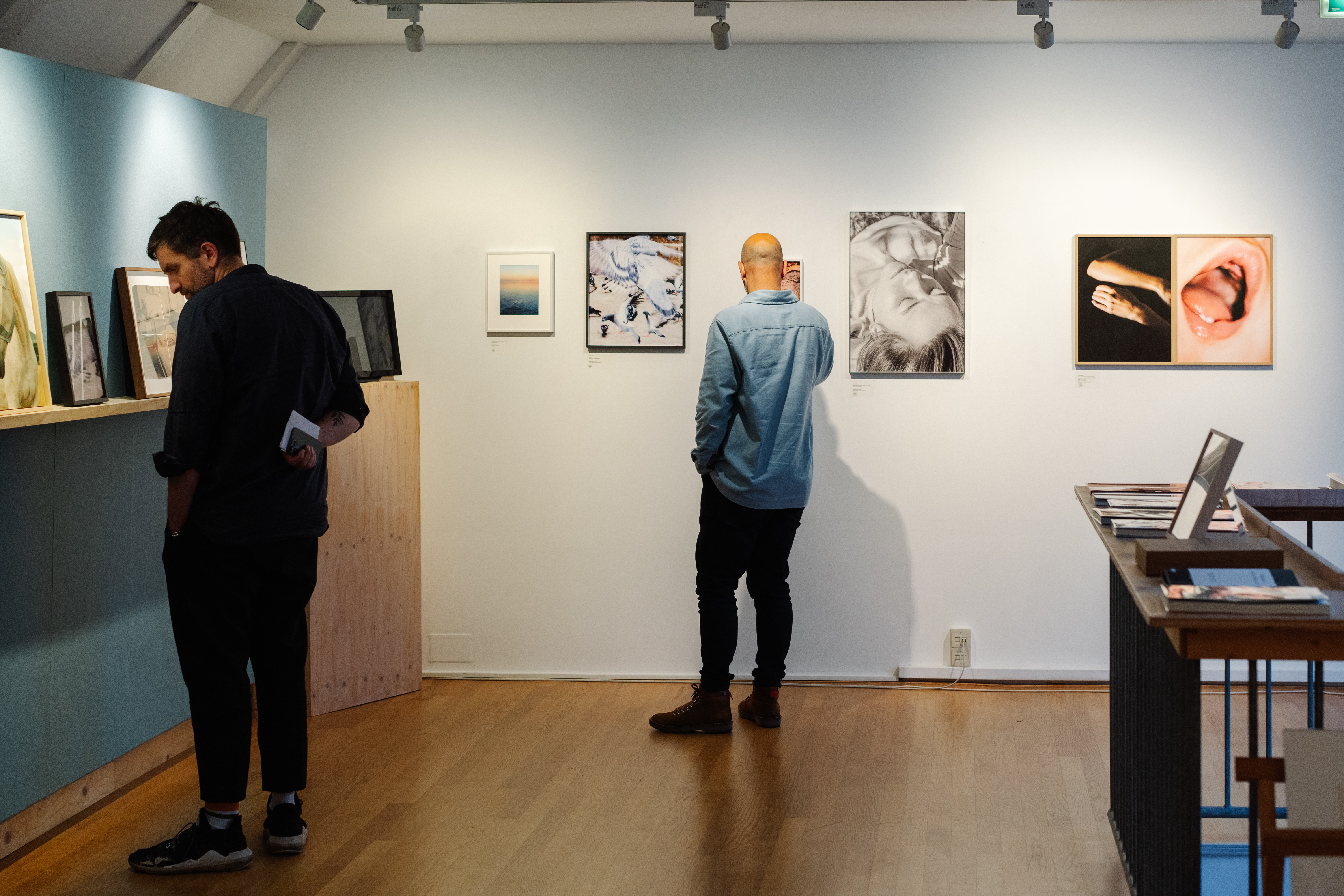 Twee mensen bekijken ingelijste foto's in een museumgalerij met witte muren, houten vloeren en railverlichting