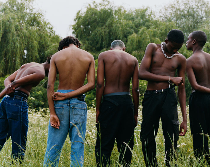 Vijf mannen zonder shirt, met hun rug naar de camera, staand in een grasveld met bomen