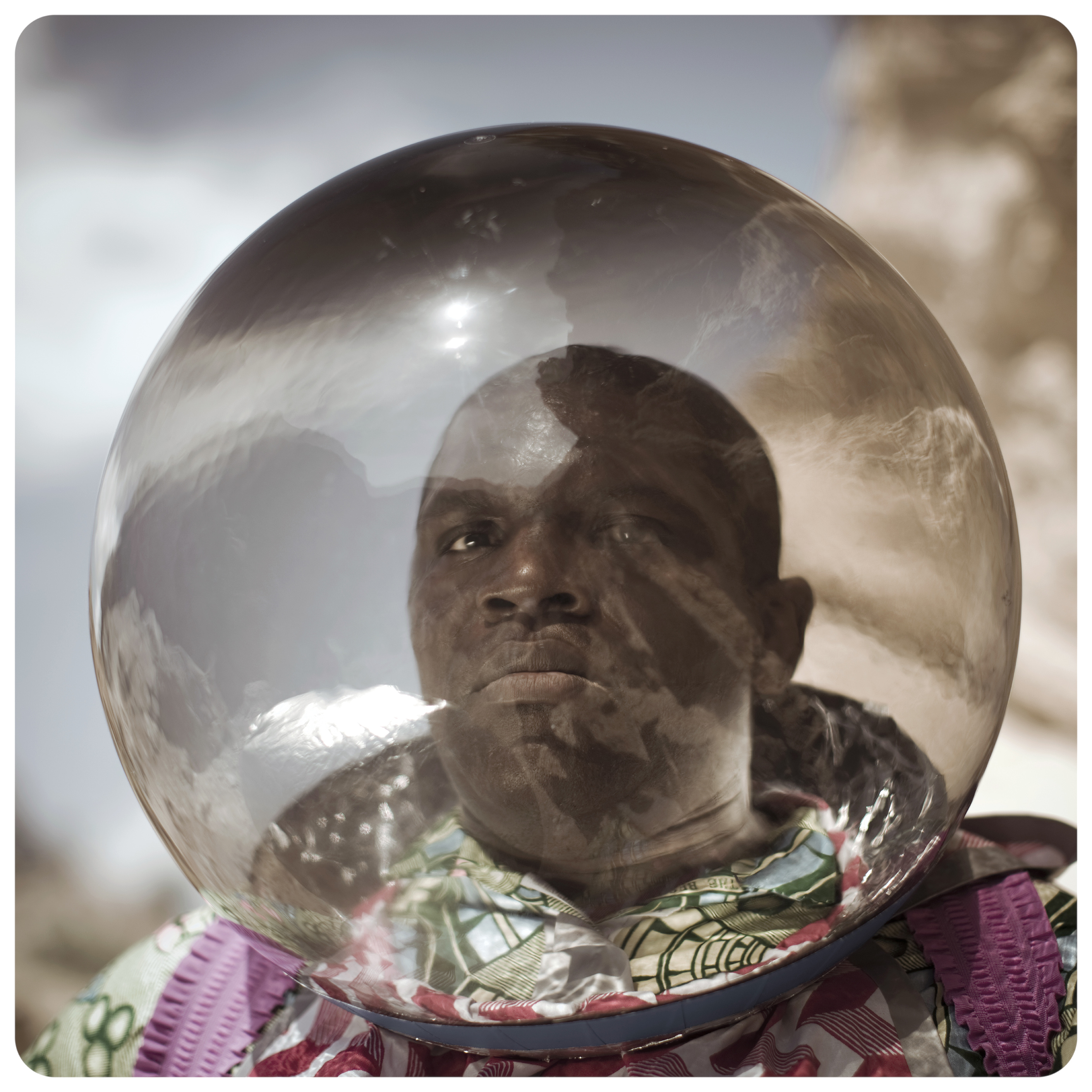 Person wearing a large transparent spherical helmet reflecting sky and clouds, colorful patterned garment, outdoor setting