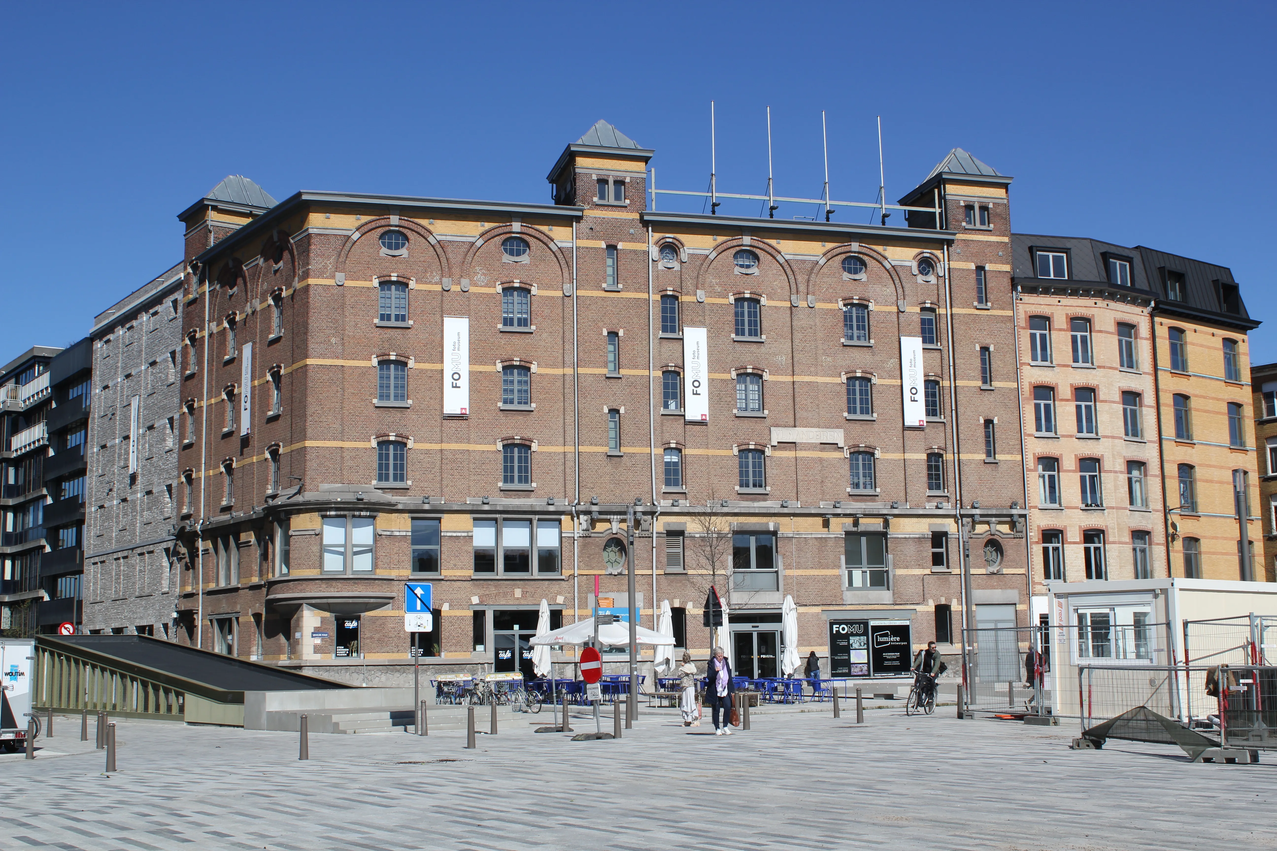 Gran edificio histórico de ladrillo con ventanas arqueadas y carteles de exposiciones blancos que cuelgan de la fachada, visto desde una plaza pavimentada bajo un cielo azul despejado