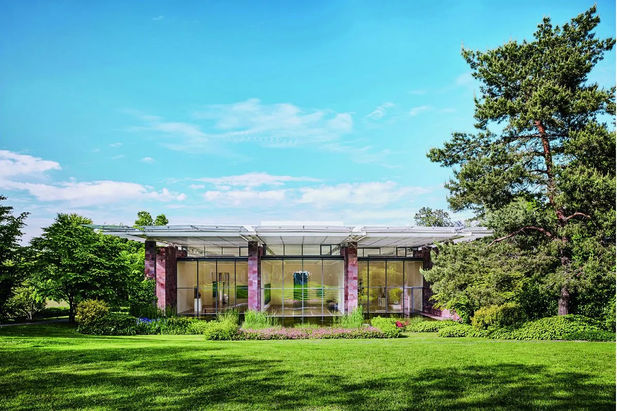 Buitenaanzicht van het museumgebouw van de Fondation Beyeler met een grote glazen gevel en stenen zuilen, omringd door een groen gazon en hoge bomen onder een blauwe lucht.
