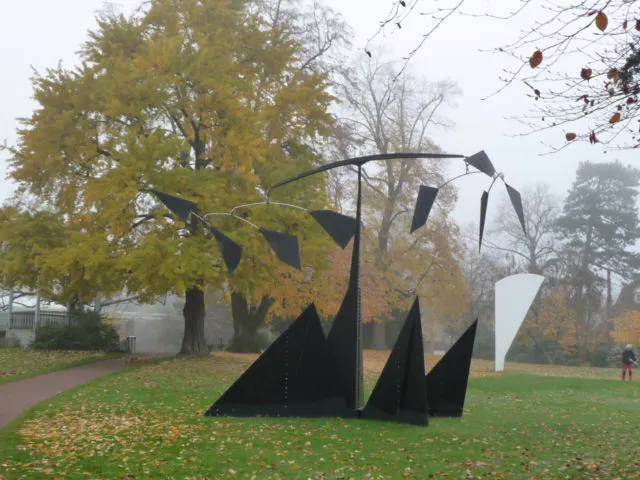 Grote zwart-witte mobiele sculptuur van Alexander Calder op een grasveld met herfstbomen en gevallen bladeren op de achtergrond