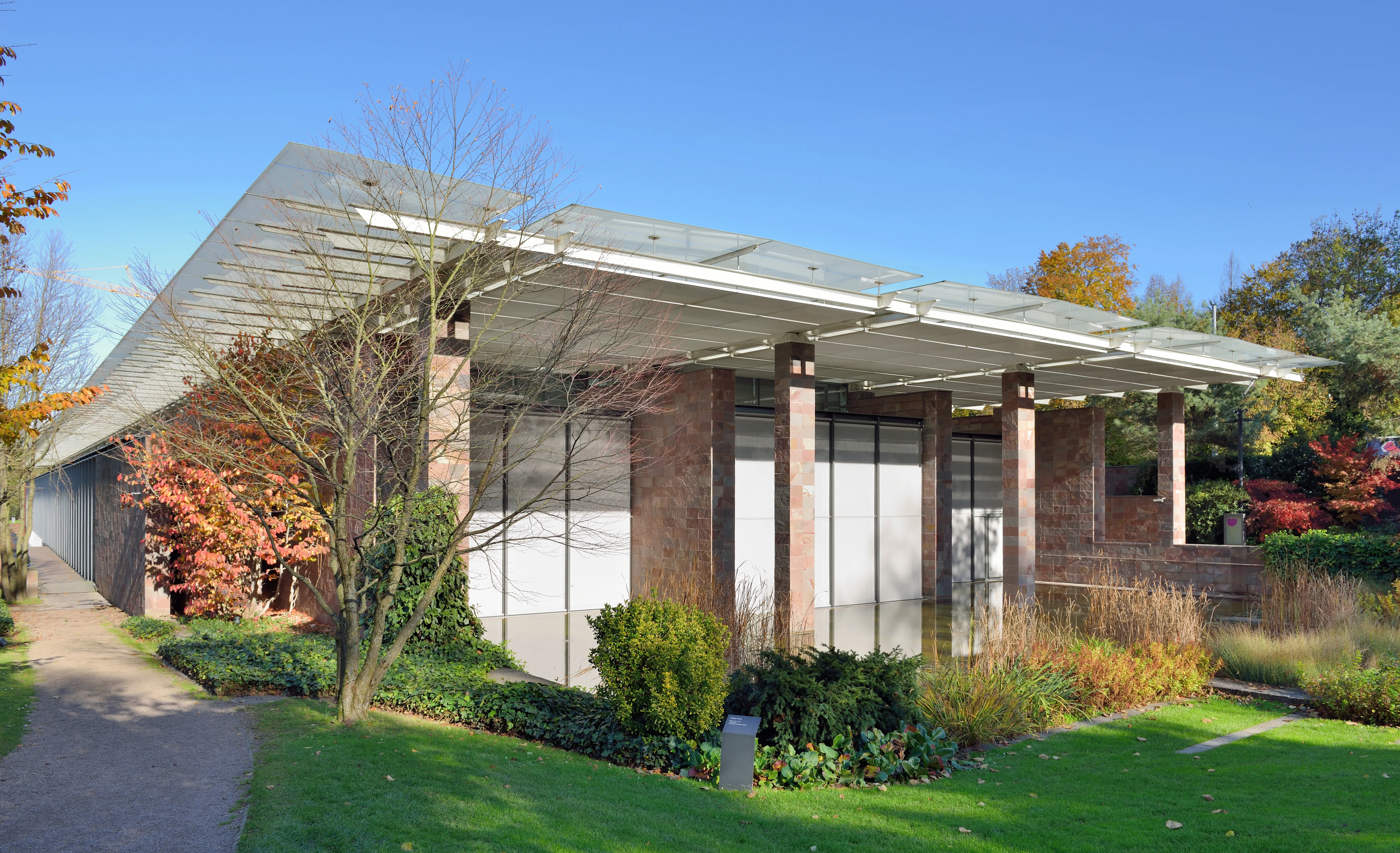 Buitenaanzicht van museum Fondation Beyeler met het karakteristieke platte glazen dak van Renzo Piano, stenen zuilen en glazen wanden, omgeven door een herfstachtig landschap.