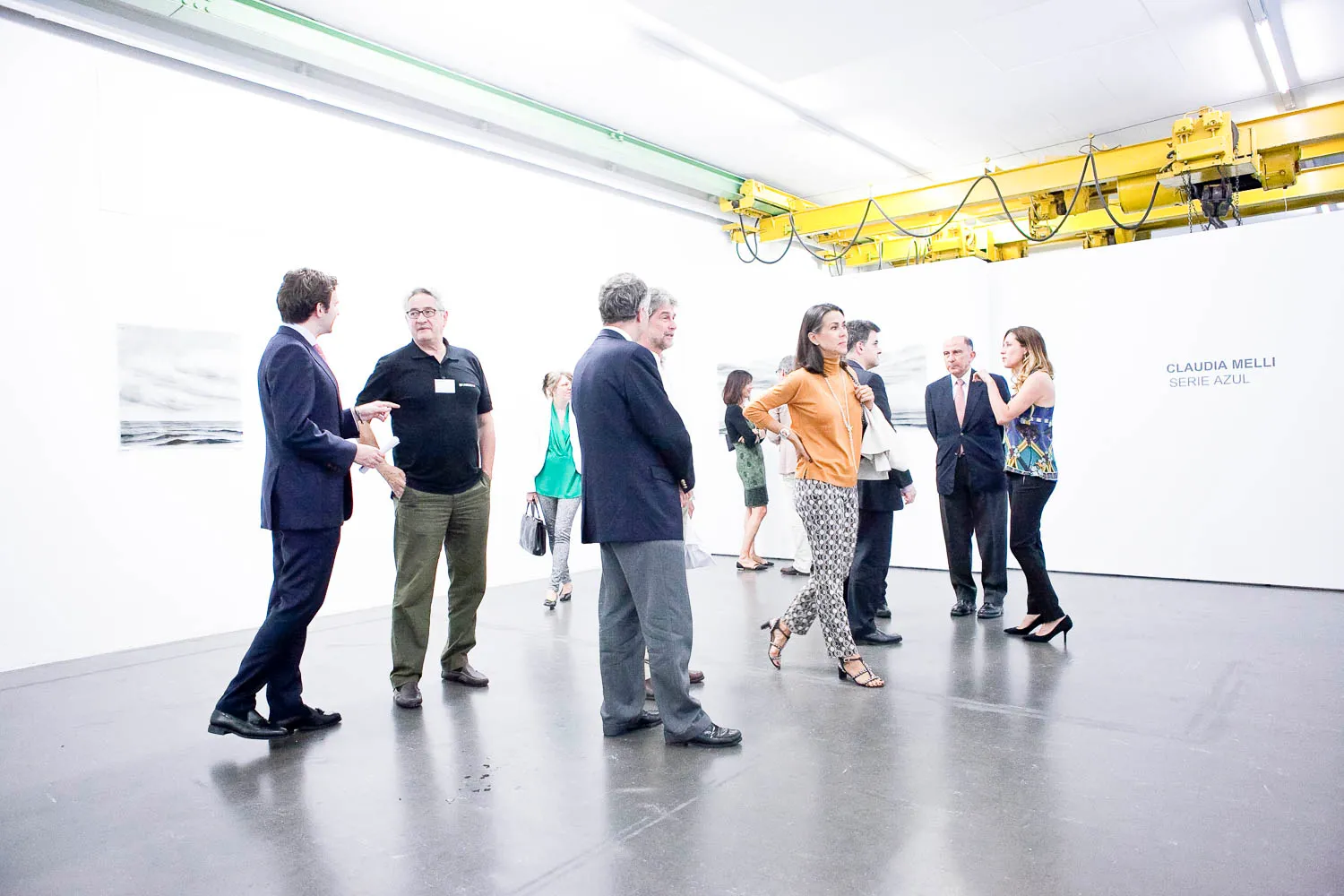 Grupo de visitantes charlando en una luminosa galería de paredes blancas con una grúa industrial amarilla en el techo y suelo de hormigón pulido.
