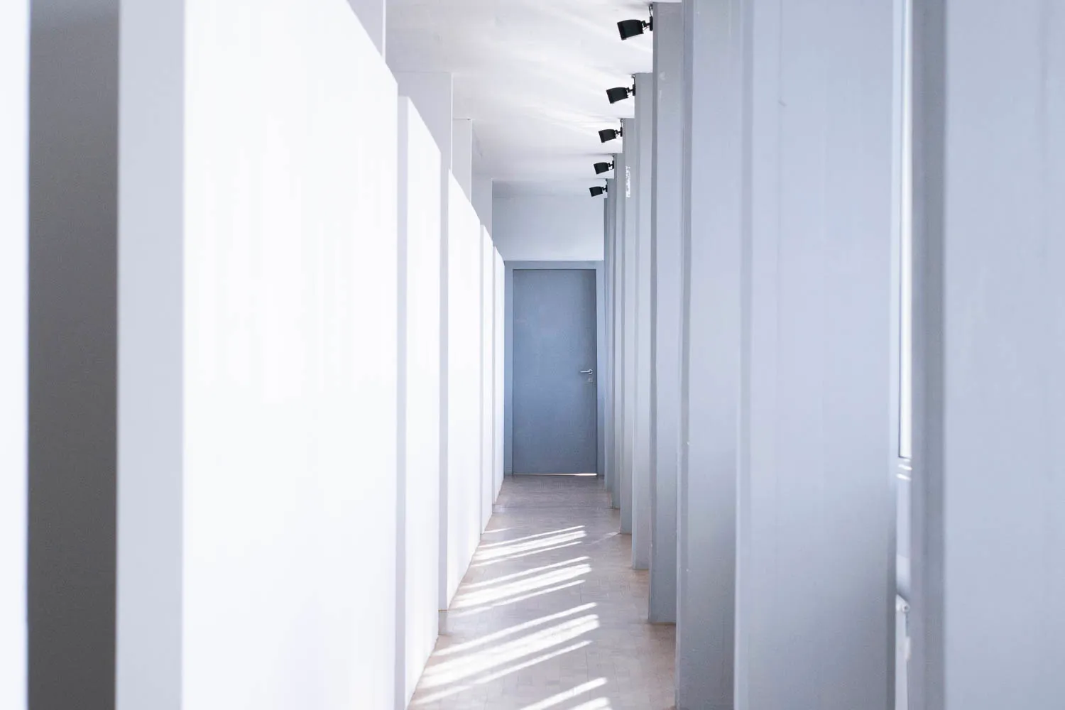 Un pasillo largo y de color blanco brillante con luz solar que proyecta sombras a rayas en el suelo, iluminación de riel a lo largo del techo y una puerta azul grisáceo visible al fondo.