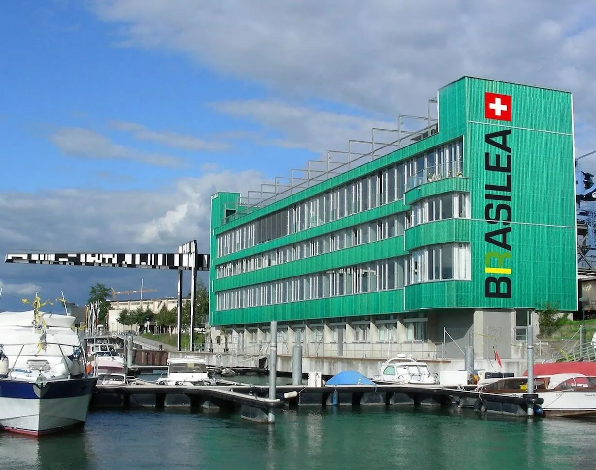 Edificio verde de varias plantas con 'BRASILEA' escrito verticalmente en la fachada junto a una cruz suiza, situado en un puerto con barcos atracados delante